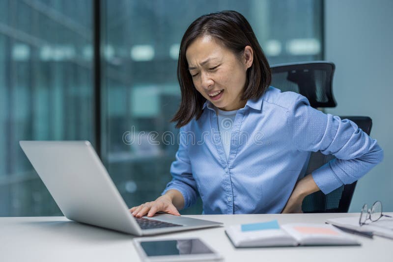 Une femme d'affaires asiatique souffle de douleur en travaillant sur son ordinateur portable souffrant de maux de dos. photo stock