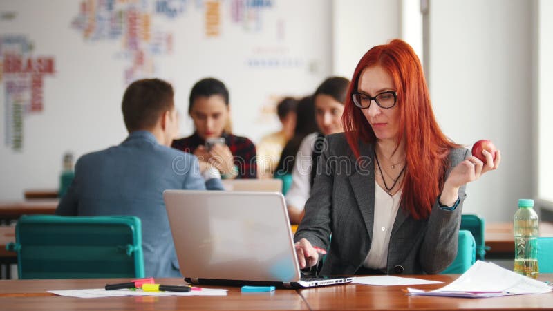 Une femme de gingembre en verres se reposant au bureau fonctionnant à côté de l'ordinateur portable et mangeant la pomme banque de vidéos