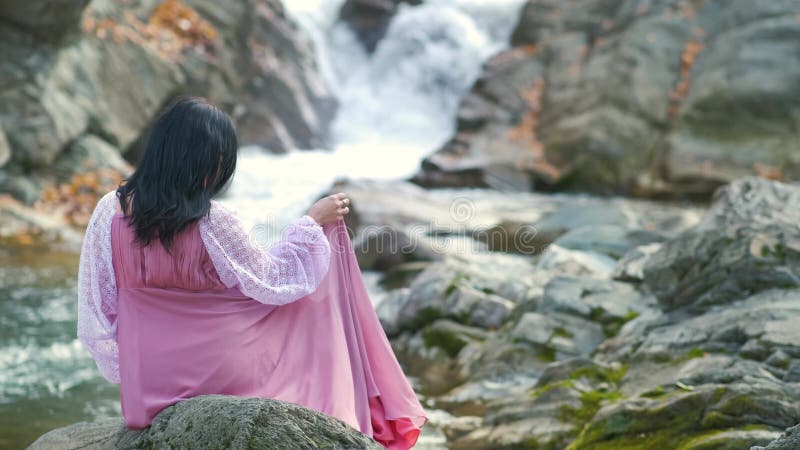 Une femme en longue robe de soirée à la mode assise sur une pierre près d'une petite rivière de montagne avec de l'eau à mouvement banque de vidéos