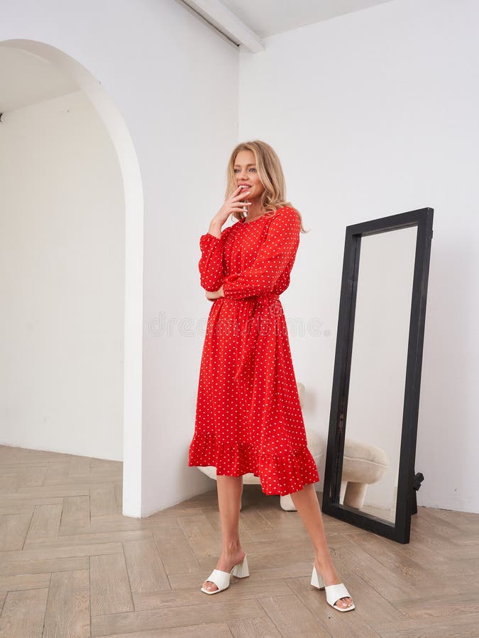Une femme en robe rouge polkadot se tient dans une pièce minimaliste. Elle porte des sandales blanches d'une main photo libre de droits