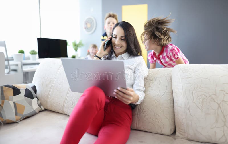 Une femme est assise sur un canapé tenant un ordinateur portable et parlant sur un smartphone à côté des enfants jouent. images stock
