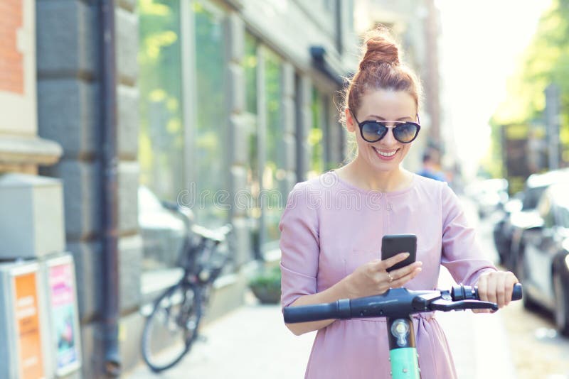 Une femme heureuse en lunettes de soleil en utilisant un smartphone et en louant un scooter électrique moderne avec une applicati photographie stock Une femme heureuse en lunettes de soleil en utilisant un smartphone et en louant un scooter électrique moderne avec une applicati photographie stock