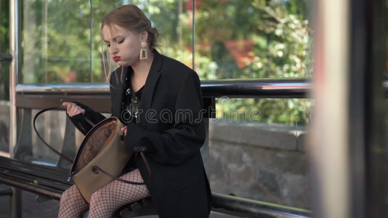 Une femme inquiète et stressée avec un sac à main vide assis sur un arrêt de bus à l'extérieur. portrait de jeune belle caucasienn banque de vidéos