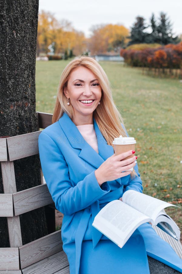 Une femme souriante est assise près d'un arbre dans un parc d'automne et tient un livre et une tasse avec une boisson chaude dans images stock Une femme souriante est assise près d'un arbre dans un parc d'automne et tient un livre et une tasse avec une boisson chaude dans images stock
