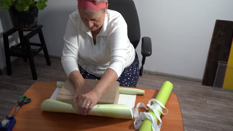 Une femme âgée enveloppe un cadeau dans du papier. Une grand-mère fait une boîte banque de vidéos