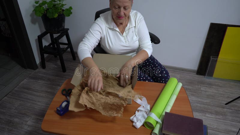 Une femme âgée enveloppe un cadeau dans du papier. Une grand-mère fait une boîte banque de vidéos