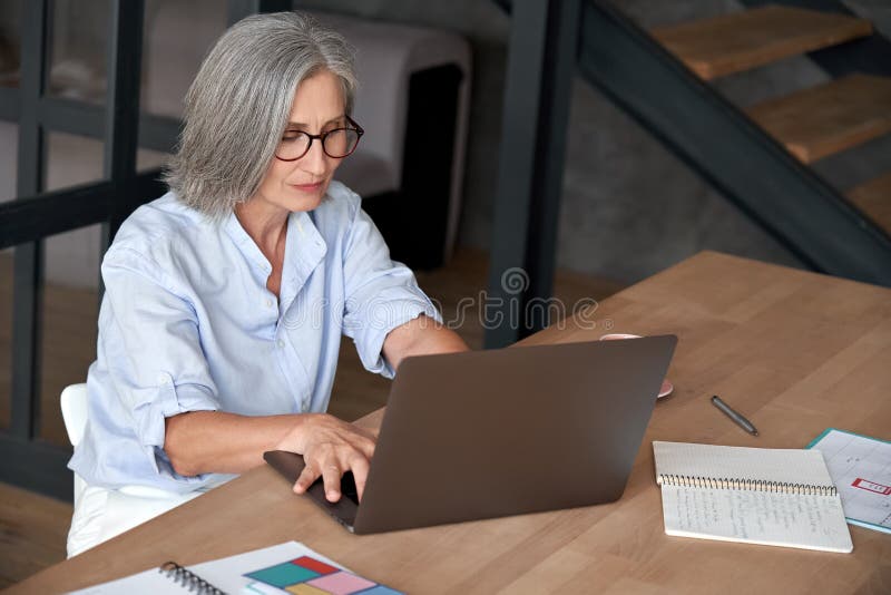 Une femme âgée moyenne mûre aînée à l'aide de l'ordinateur portable assis au bureau photos stock