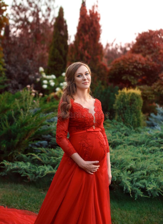 Une femme élégante et enceinte marche dans un beau jardin dans une robe rouge luxueuse et chère avec un long train artistique photo libre de droits Une femme élégante et enceinte marche dans un beau jardin dans une robe rouge luxueuse et chère avec un long train artistique photo libre de droits