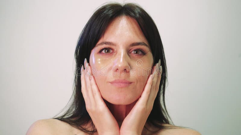 Une fille aux cheveux noirs applique des cosmétiques à son visage. jeune femme aux patchs cosmétiques sous ses yeux. une closeup g banque de vidéos