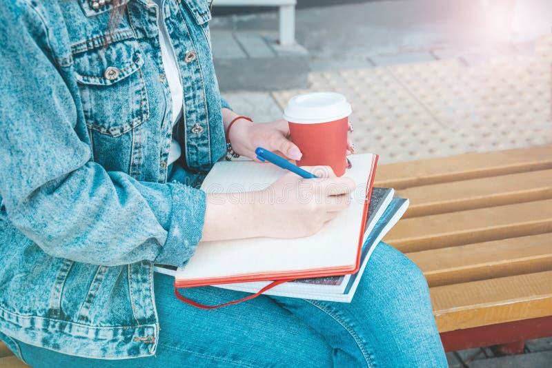 Une fille dans des vêtements de jeans s'assied sur un banc, tient des carnets et écrit Sur la rue, elle écrit dans un carnet, étu image stock Une fille dans des vêtements de jeans s'assied sur un banc, tient des carnets et écrit Sur la rue, elle écrit dans un carnet, étu image stock