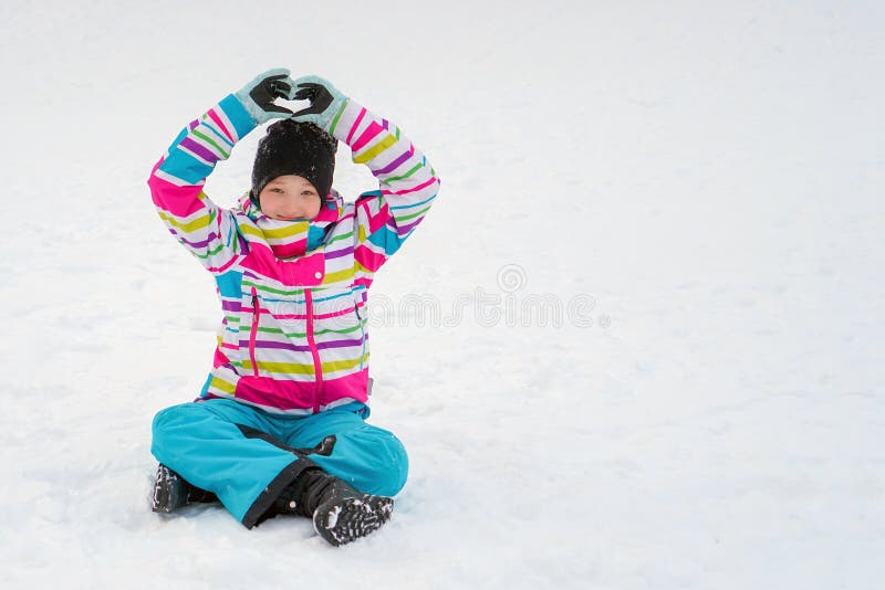 Une fille dans un pullover de ski coloré lumineux dans une veste lumineuse s'assied, sur le bonheur et montre le coeur avec ses m photo libre de droits Une fille dans un pullover de ski coloré lumineux dans une veste lumineuse s'assied, sur le bonheur et montre le coeur avec ses m photo libre de droits