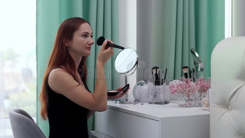 Une fille en robe de soirée lui applique de la poudre au visage. Jeune femme attrayante faisant du maquillage derrière une coiffeu banque de vidéos