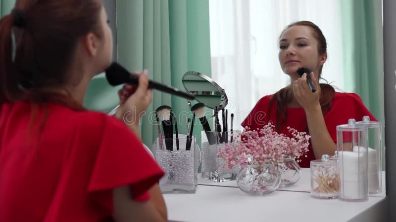 Une fille en robe de soirée lui applique de la poudre au visage. Jeune femme attrayante faisant du maquillage derrière une coiffeu banque de vidéos