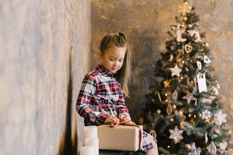 Une fillette de six ans vêtue d'une robe de plaid à l'arrière d'un arbre de Noël ouvre son cadeau de nouvelle année images stock Une fillette de six ans vêtue d'une robe de plaid à l'arrière d'un arbre de Noël ouvre son cadeau de nouvelle année images stock
