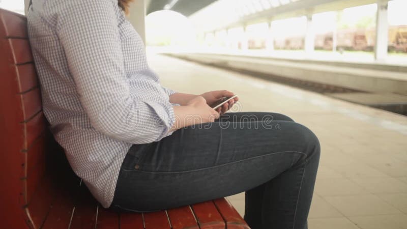 Une jeune femme assise sur un banc. gare. la jeune fille écrit un message sur le téléphone utilise internet en attendant banque de vidéos