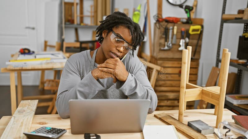 Une jeune femme noire avec lunettes et poignards s'assoit avec soin à un bureau en bois dans un atelier de menuiserie images libres de droits