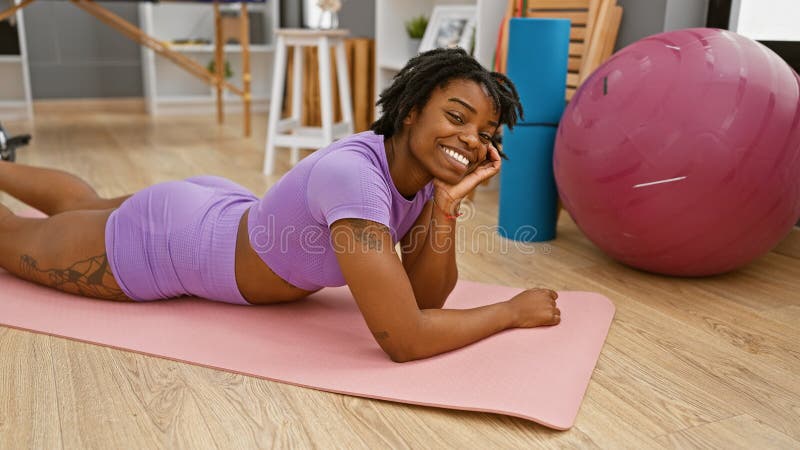 Une jeune femme noire souriante aux cadenas allongés sur un tapis de yoga à l'intérieur près d'une balle de fitness image stock