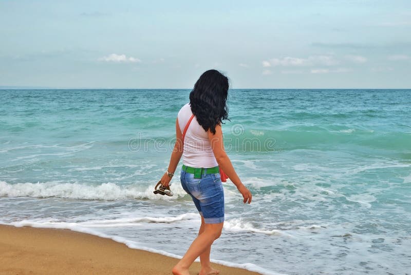 Une jeune fille dans un T-shirt blanc marche le long du sable en mer brune sur le rivage de la mer azurée en Bulgarie photographie stock libre de droits Une jeune fille dans un T-shirt blanc marche le long du sable en mer brune sur le rivage de la mer azurée en Bulgarie photographie stock libre de droits