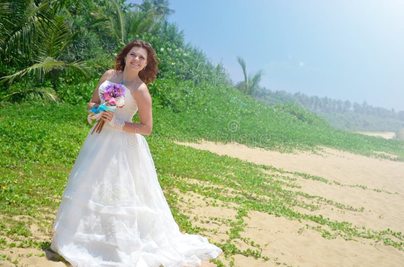 Une jeune jeune mariée dans une robe bien aérée blanche se tient avec un bouquet des lotus fille souriant sur une plage tropicale photos libres de droits Une jeune jeune mariée dans une robe bien aérée blanche se tient avec un bouquet des lotus fille souriant sur une plage tropicale photos libres de droits
