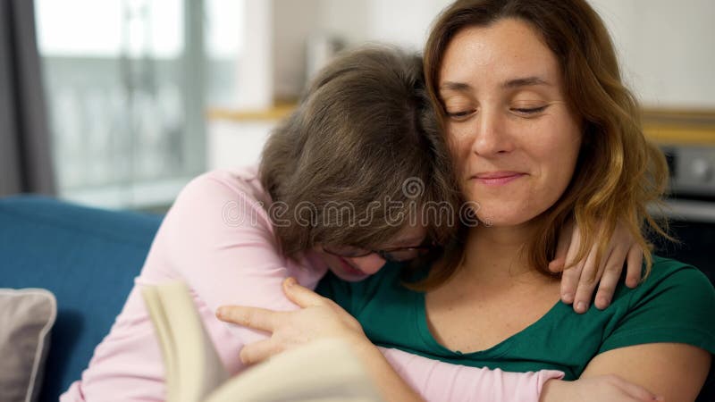 Une jeune mère lit un livre à sa fille avec le syndrome du bas pendant qu'ils sont assis sur le canapé dans la pièce banque de vidéos