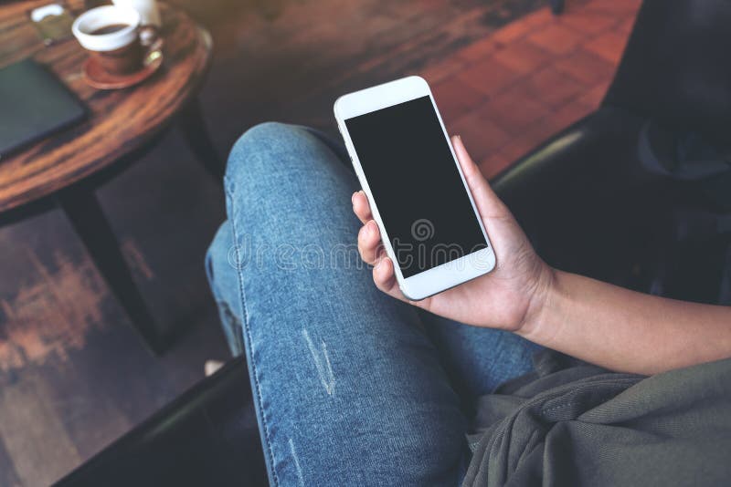 Top view mockup image of a woman`s hand holding white mobile phone with blank black desktop screen on thigh while sitting in cafe. Top view mockup image of a woman`s hand holding white mobile phone with blank black desktop screen on thigh while sitting in cafe