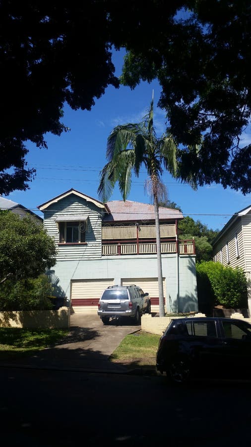 Une maison plus ancienne de Queenslander avec le garage de bloc de ciment dessous et la voiture a garé dans l'allée encadrée par photographie stock Une maison plus ancienne de Queenslander avec le garage de bloc de ciment dessous et la voiture a garé dans l'allée encadrée par photographie stock