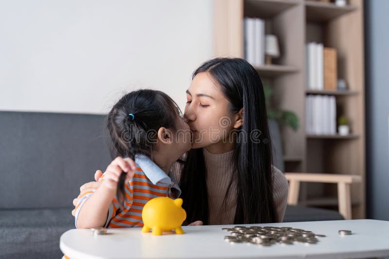Une mère diversifiée partageant un baiser amoureux avec sa fille tout en économisant de l'argent avec une tirelire jaune et des pi photo libre de droits