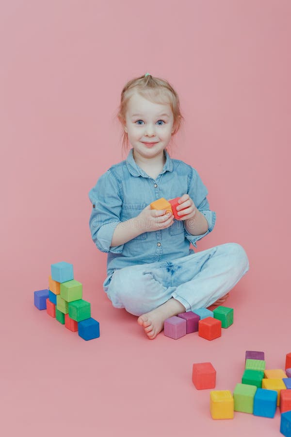 Une petite fille de charme dans des vêtements de denim sur un fond rose joue avec les cubes colorés en bois images stock Une petite fille de charme dans des vêtements de denim sur un fond rose joue avec les cubes colorés en bois images stock