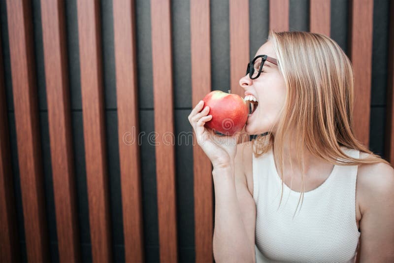 Une photo de fille blonde mordant un morceau de pomme rouge Elle apprécie le moment D'isolement sur le fond rayé photo stock Une photo de fille blonde mordant un morceau de pomme rouge Elle apprécie le moment D'isolement sur le fond rayé photo stock