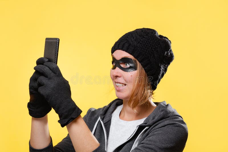 A female hacker in a black hat, gloves and glasses holds a mobile phone in her hands, trying to hack into a database. Yellow background. The concept of cybercrime and hacking. A female hacker in a black hat, gloves and glasses holds a mobile phone in her hands, trying to hack into a database. Yellow background. The concept of cybercrime and hacking.