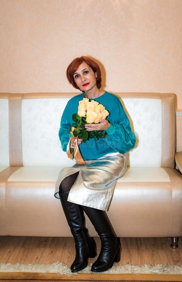 Une russe de 48 ans en bonne santé avec des cheveux rouges vibrants et des vêtements élégants assis sur un canapé avec un bouquet image libre de droits