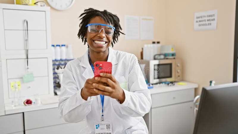 Une scientifique joyeuse avec des poignées de redirection à l'aide d'un smartphone dans un laboratoire portant un manteau blanc et images stock