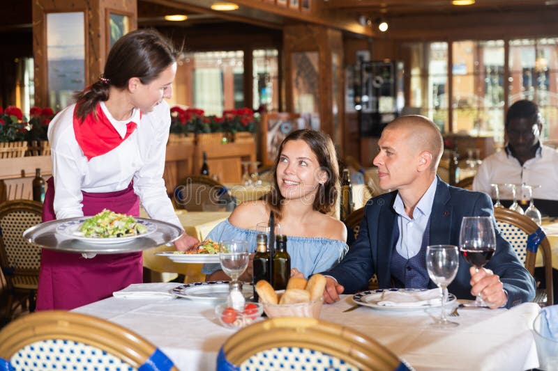 Smiling waitress serving food for positive couple at restaurant, putting plate with salad on table. Smiling waitress serving food for positive couple at restaurant, putting plate with salad on table