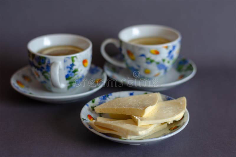 A saucer with sliced cheese and two cups of tea with lemon on a gray background. Ceramic tableware decorated with camomiles. Teatime. Side view from an angle. A saucer with sliced cheese and two cups of tea with lemon on a gray background. Ceramic tableware decorated with camomiles. Teatime. Side view from an angle