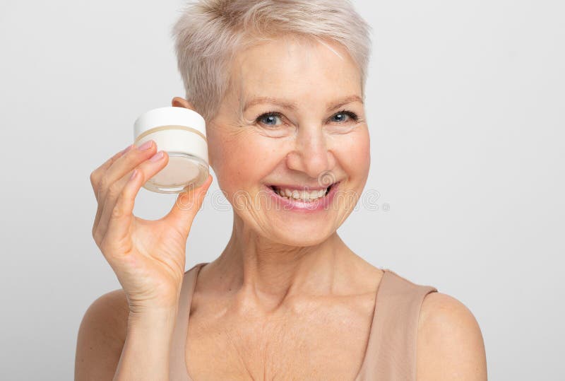 Une vieille dame charmante avec une courte coiffure sourit et tient un bocal de crème dans les mains. routine beauté. image stock