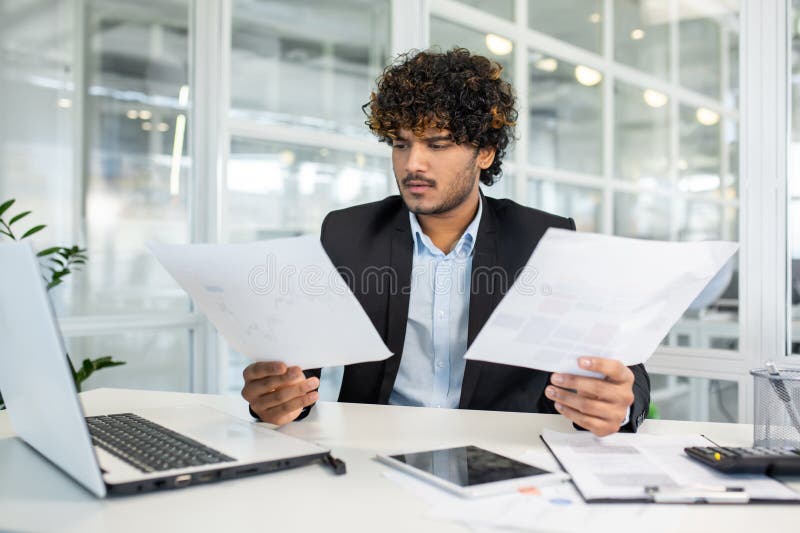 Focused young businessman with curly hair wearing a suit examining multiple documents and reports while sitting at a modern office desk. Focused young businessman with curly hair wearing a suit examining multiple documents and reports while sitting at a modern office desk.