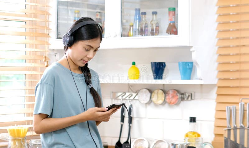Ung asiatisk kvinna med hörlurar som lyssnar till musik från hemmastadd kökbakgrund för mobiltelefon, folk och teknologilivsstil fotografering för bildbyråer Ung asiatisk kvinna med hörlurar som lyssnar till musik från hemmastadd kökbakgrund för mobiltelefon, folk och teknologilivsstil fotografering för bildbyråer