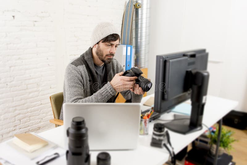 Ung attraktiv pressfotograf som rymmer den fotografiska kameran som beskådar hans arbete på redaktörkontorsskrivbordet royaltyfria bilder Ung attraktiv pressfotograf som rymmer den fotografiska kameran som beskådar hans arbete på redaktörkontorsskrivbordet royaltyfria bilder