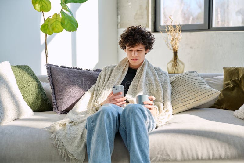 Ung avslappnad kille under varm filt med mycket smartphone sittande eller sofa hemma arkivfoto