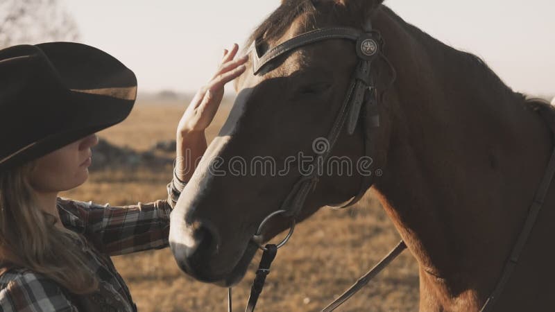 Ung blond flicka med långt hår i cowboyhatt som slår och kramar en häst
