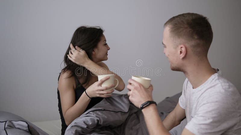 Ung caucasian man i den vita T-skjortan att komma med kaffe i säng till hans älskade kvinna Lycka som känner sig bra romantisk mo