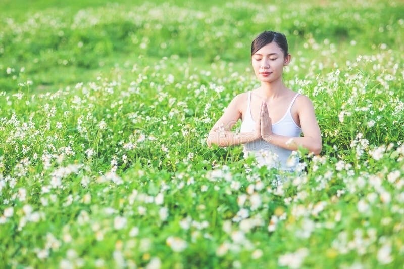 Ung dam som gör yogaövning i grönt fält med litet utomhus- område för vita blommor som visar fridsam stillhet i meditationmening royaltyfri bild Ung dam som gör yogaövning i grönt fält med litet utomhus- område för vita blommor som visar fridsam stillhet i meditationmening royaltyfri bild