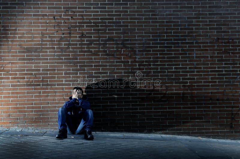 Young desperate man who lost job abandoned and lost in depression sitting on ground street corner against brick wall suffering emotional pain, crying alone in grunge lighting. Young desperate man who lost job abandoned and lost in depression sitting on ground street corner against brick wall suffering emotional pain, crying alone in grunge lighting