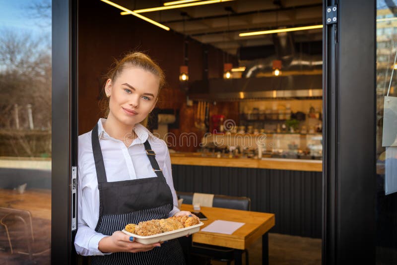 Portrait of smiling owner standing at restaurant door holding baked meatballs. Young entrepreneur woman at small family business restaurant leaning at entrance door inviting people to taste food. Portrait of smiling owner standing at restaurant door holding baked meatballs. Young entrepreneur woman at small family business restaurant leaning at entrance door inviting people to taste food