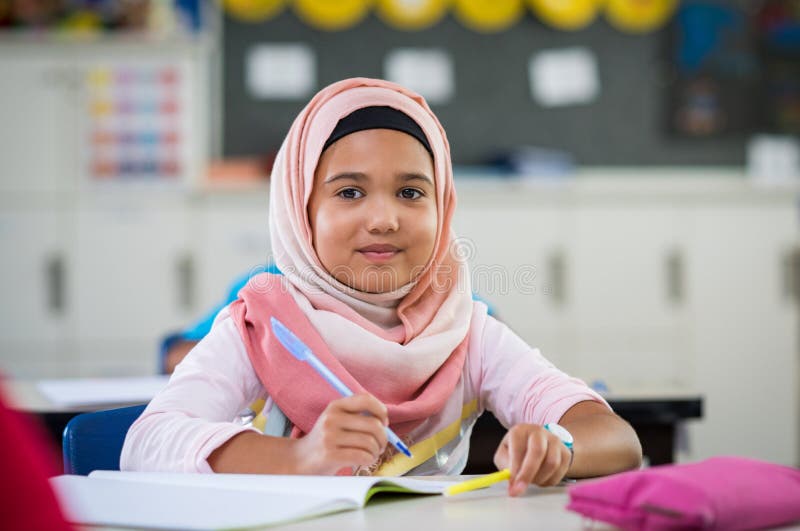 Happy young smiling girl wearing hijab and looking at camera while sitting at desk with book and pen in hand. Elementary muslim schoolgirl writing notes in classroom. Portrait of arab school girl in chador. Happy young smiling girl wearing hijab and looking at camera while sitting at desk with book and pen in hand. Elementary muslim schoolgirl writing notes in classroom. Portrait of arab school girl in chador.