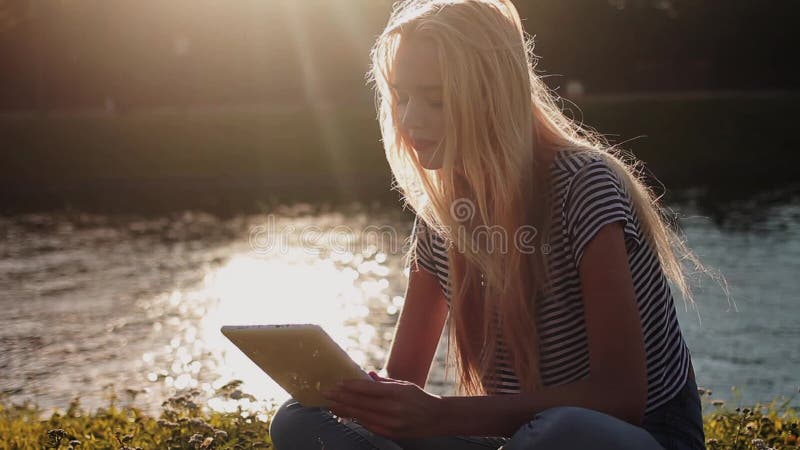 Ung flicka med minnestavlaPC:n som sitter på det gröna gräset nära floden
