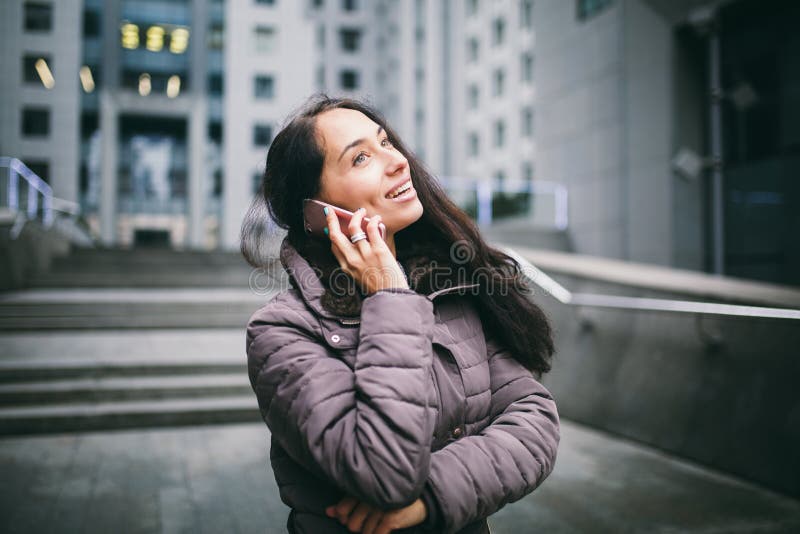 Ung flicka som talar på mobiltelefonen i borggårdaffärsmitt flicka med det långa iklädda vinteromslaget för mörkt hår i kallt väd. Ung flicka som talar på mobiltelefonen i borggårdaffärsmitt flickan med det långa iklädda vinteromslaget för mörkt hår i kallt väder talar på telefonen på bakgrundsbyggnader som göras av exponeringsglas och betong