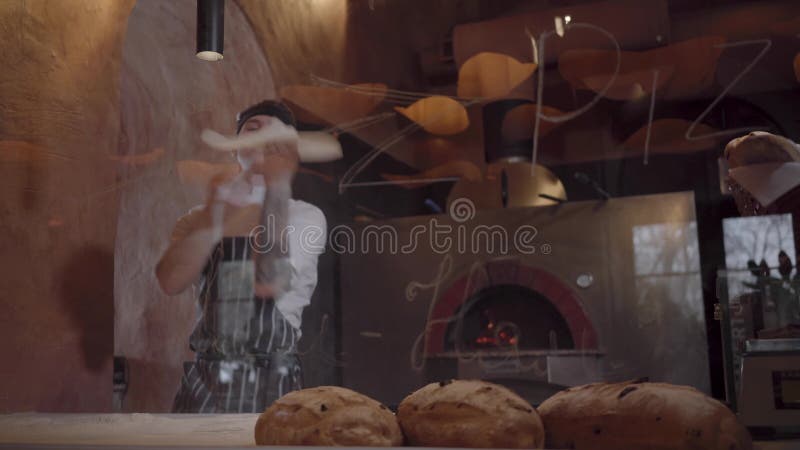 Ung grabb i kocklikformign som rotera och kastar pizzadeg som kastar det upp bak exponeringsglas i restaurangkök skillful