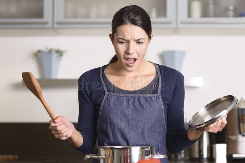 Young housewife having a calamity in the kitchen reacting in shock and horror as she lifts the lid on the saucepan on the stove to view the contents as she cooks dinner. Young housewife having a calamity in the kitchen reacting in shock and horror as she lifts the lid on the saucepan on the stove to view the contents as she cooks dinner