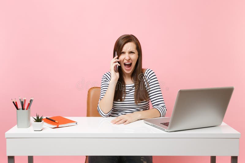 Young angry woman screaming talking on mobile phone while sitting and working on project at office with pc laptop isolated on pastel pink background. Achievement business career concept. Copy space. Young angry woman screaming talking on mobile phone while sitting and working on project at office with pc laptop isolated on pastel pink background. Achievement business career concept. Copy space